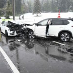 zwei weiße frontal kollidierte, im Vorderbereich stark beschädigte Autos. Im Hintergrund ein Rettungswagen, neben der Strasse liegt Schnee