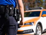 Symbolfoto: angeschnittene Ansicht eines Polizisten, der auf ein Polizeiauto im Hintergrund schaut