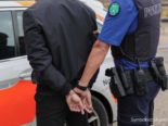 Mann in schwarzer Kleidung wird in Handschellen von Polizist zu Polizeiwagen geführt